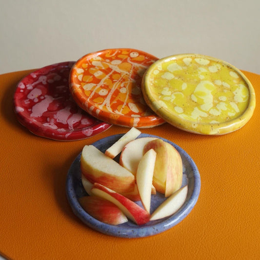 Four small handmade ceramic plates in red, orange, yellow, and blue, arranged on an orange surface with apple slices on one plate.