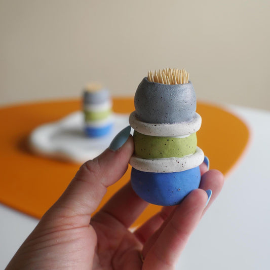 A hand holds a small ceramic toothbrush holder shaped like stacked dessert scoops in gray, white, green, and blue, with toothpicks sticking out from the top. A second matching holder is blurred in the background on an orange surface against a beige wall.