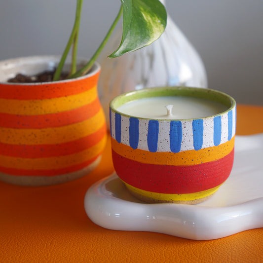 Colorful striped candle in a decorative holder with a plant and vase in the background.