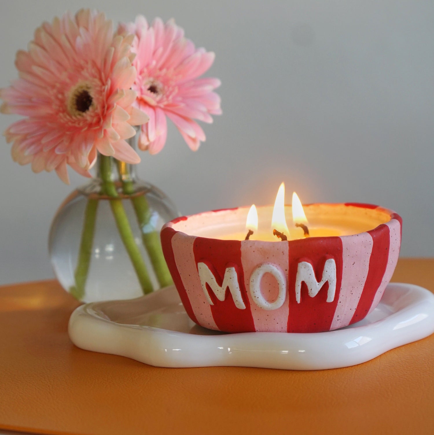 3 wick ceramic candle with red & white stripes and "MOM" in the center. Candle is burning on an orange table with pink flowers in the background