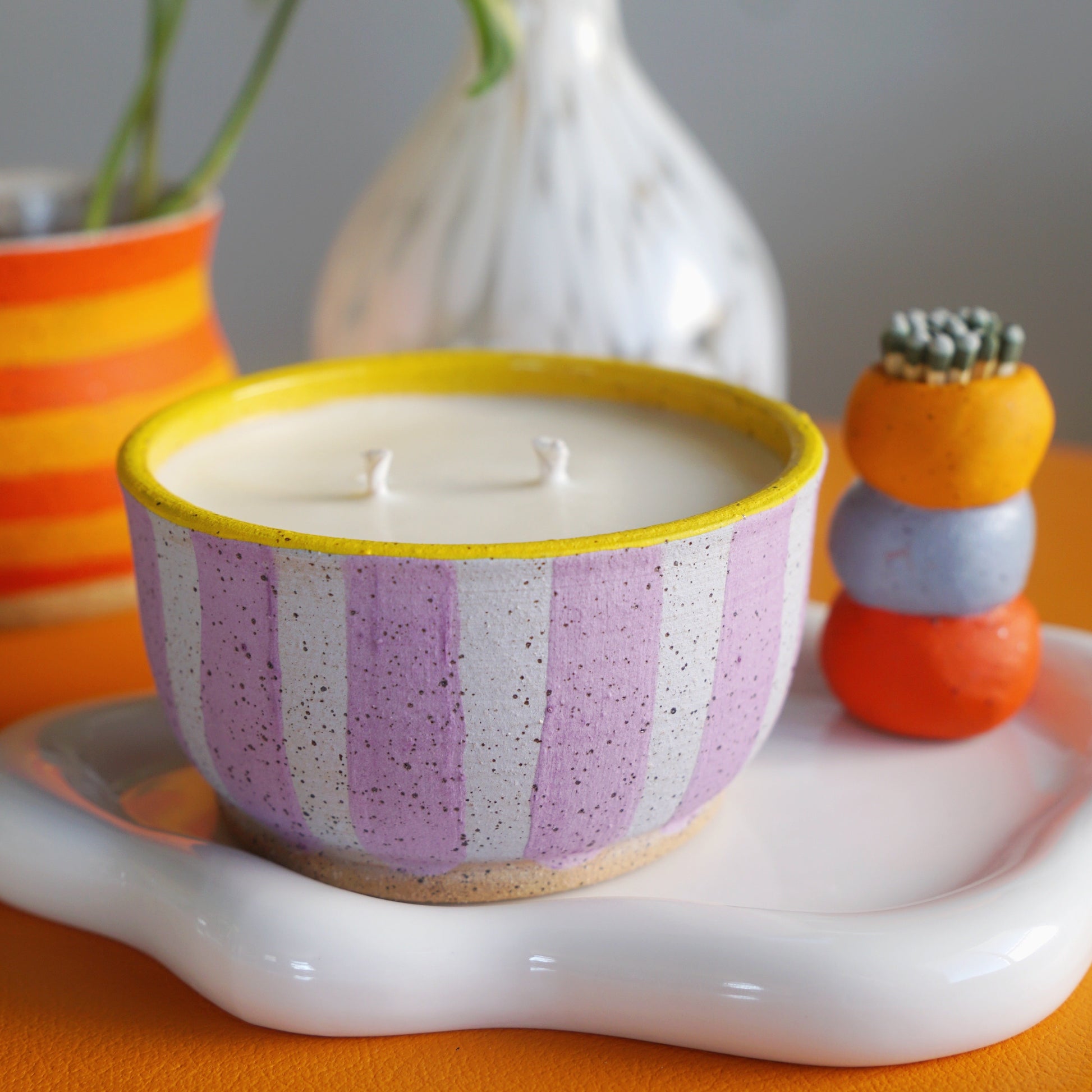 Striped candle in a decorative bowl on a white tray with a plant and small stack of colorful items in the background.