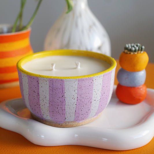 Striped candle in a decorative bowl on a white tray with a plant and small stack of colorful items in the background.