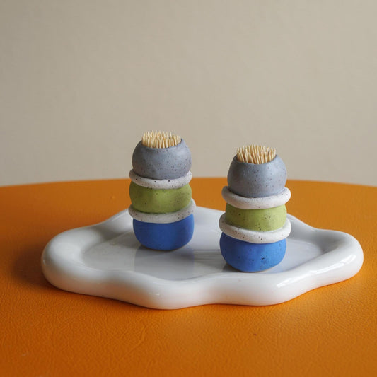 Two small handmade ceramic cactus-like figurines with stacked blue, green, and gray bodies and toothpick tops, displayed on a white tray over an orange surface against a beige background.