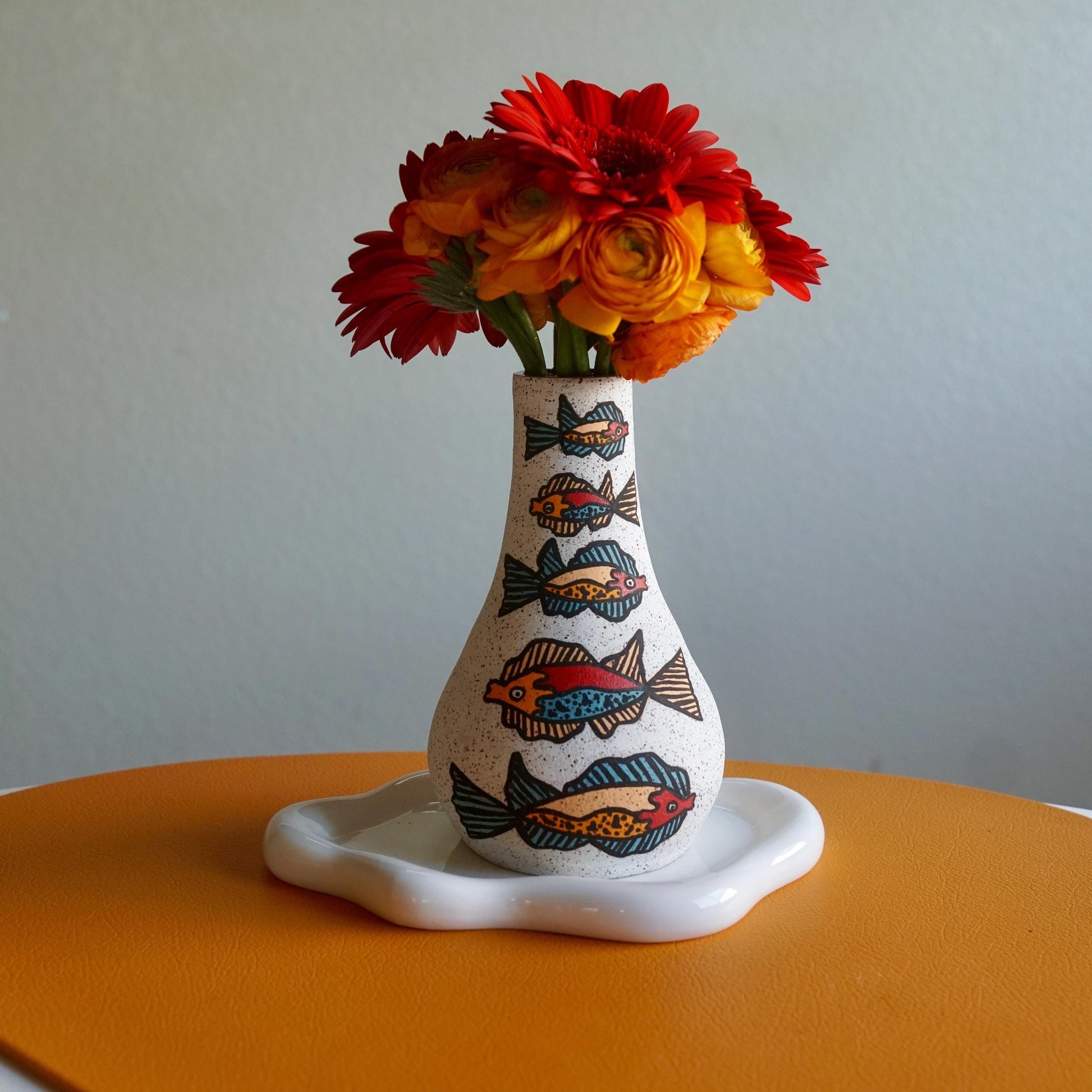 Decorative vase with fish patterns on a white base, containing red and orange flowers against a plain background.