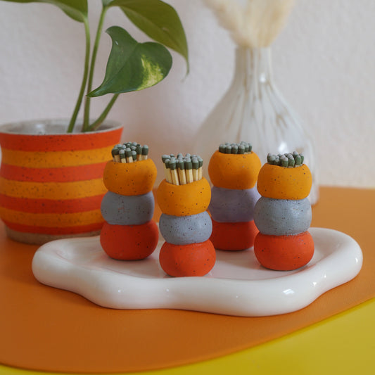 Colorful bubble shaped match holders on a white base with a plant and vase in the background.