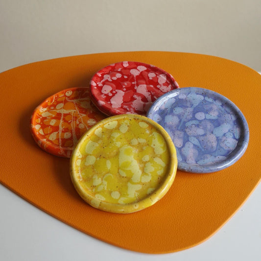 Four small round ceramic dishes in yellow, orange, red, and blue with white marbled patterns, arranged on an orange placemat against a neutral beige background.
