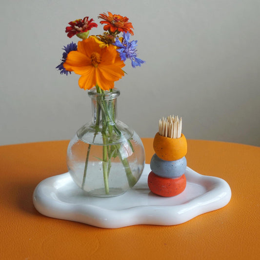 A white ceramic tray with an organic wavy edge sits on an orange surface, holding a clear glass bottle vase with colorful flowers and a small stack of three painted toothpick holders in orange, blue-gray, and red.