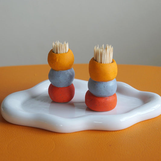 Two small toothpick holders made of stacked matte ceramic-like spheres in bright orange, light orange and purple, each holding a few wooden toothpicks, displayed on a white tray against a warm orange surface and gray background.