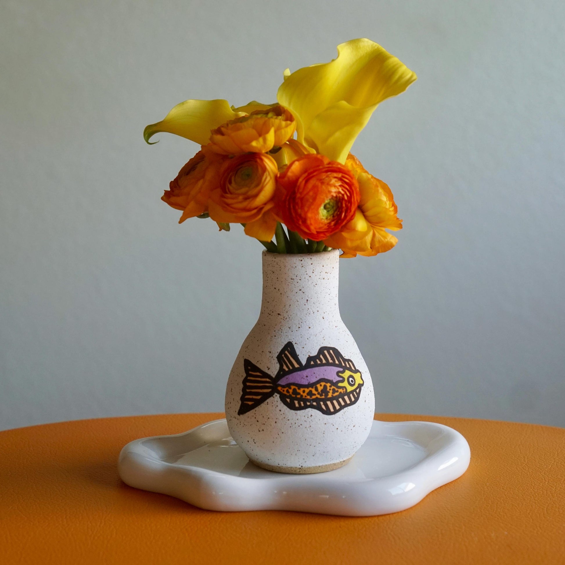 White vase with fish design holding flowers on a white plate against a gray background