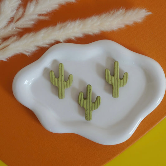 Three green cactus-shaped ceramic magnets on a white dish with pampas grass against an orange and yellow background.