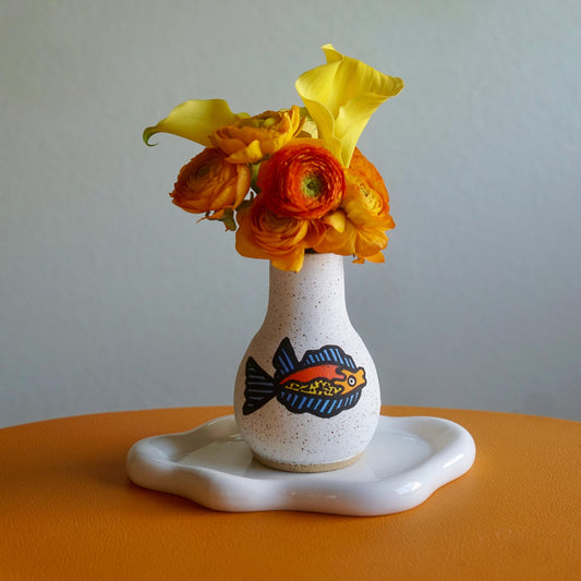 White vase with fish design on a stand holding yellow and orange flowers against a plain background