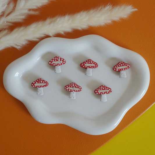Set of red mushroom-shaped magnets on a white dish with decorative pampas grass on an orange and yellow background.