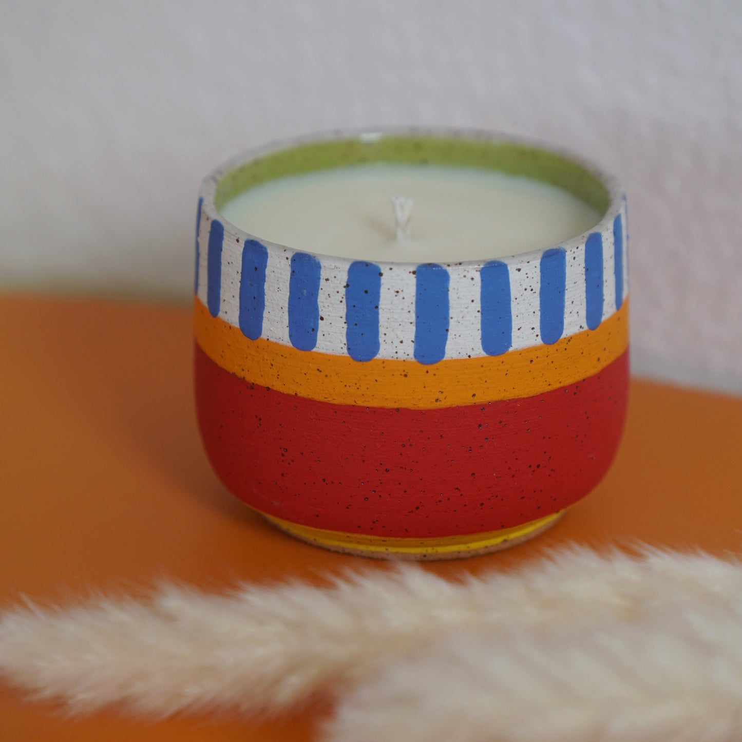 Candle in a colorful striped container on an orange surface with white pampas grass.