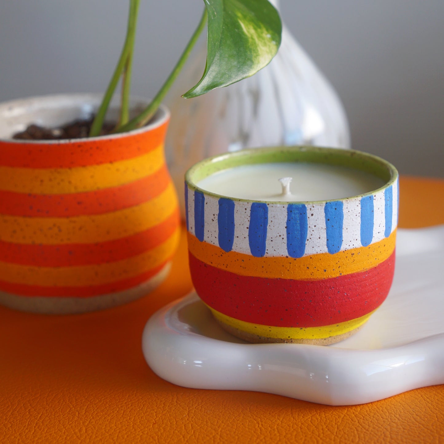 Colorful striped candle in a decorative holder with a plant and vase in the background.