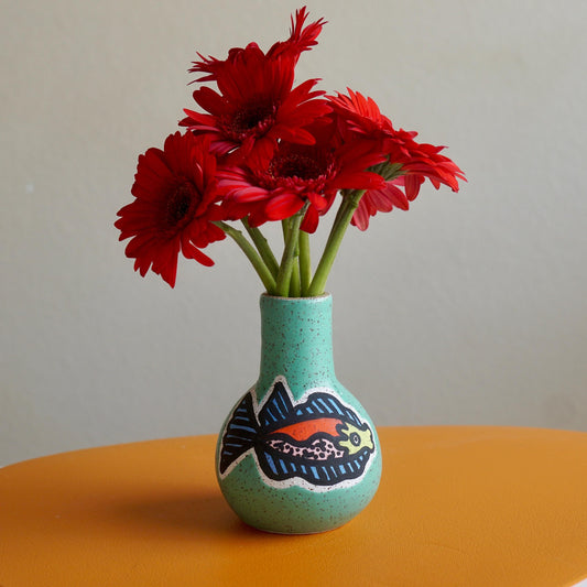 Turquoise vase with fish design containing red flowers on a yellow surface.