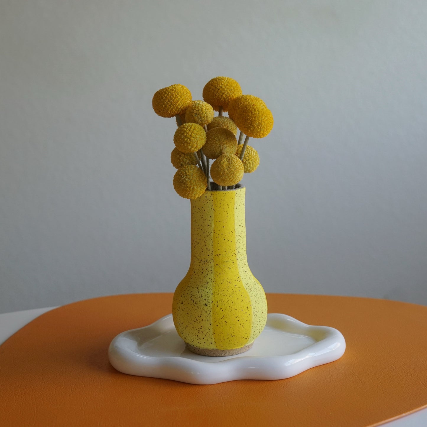 Yellow textured vase with dried yellow flowers on a white surface with an orange mat