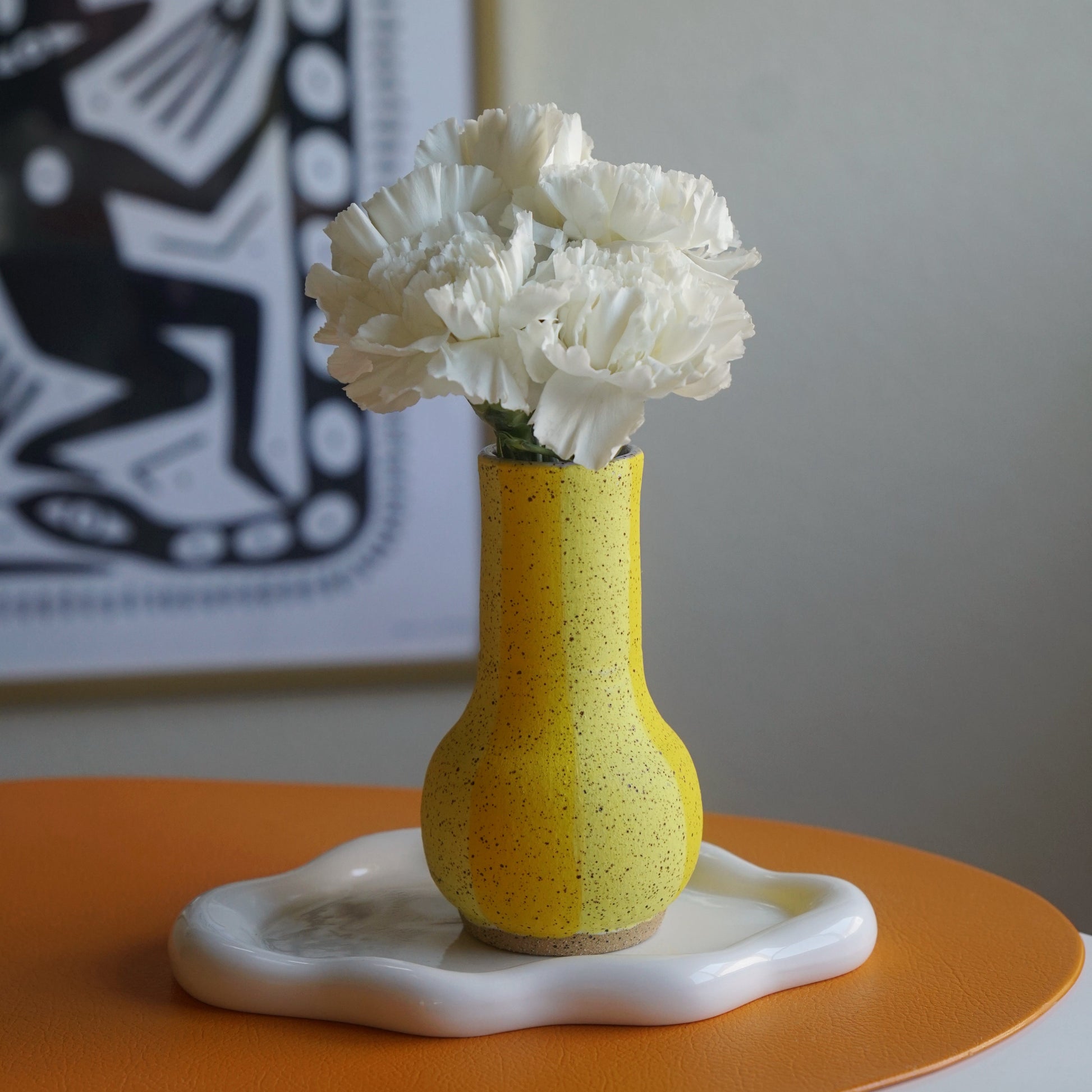Yellow vase with white flowers on an orange surface, with a abstract painting in the background.