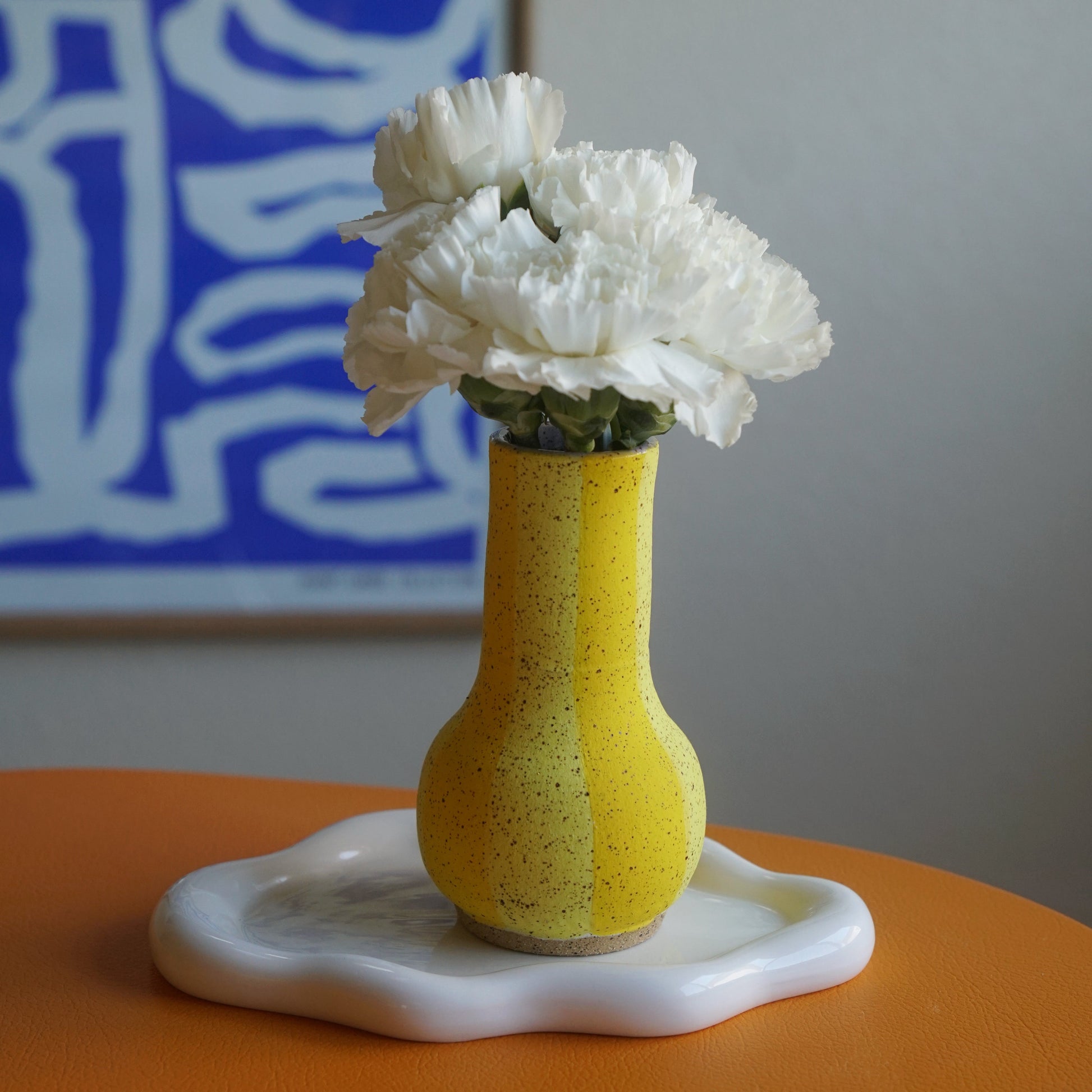 Yellow vase with white flowers on an orange surface, with a blue abstract painting in the background.