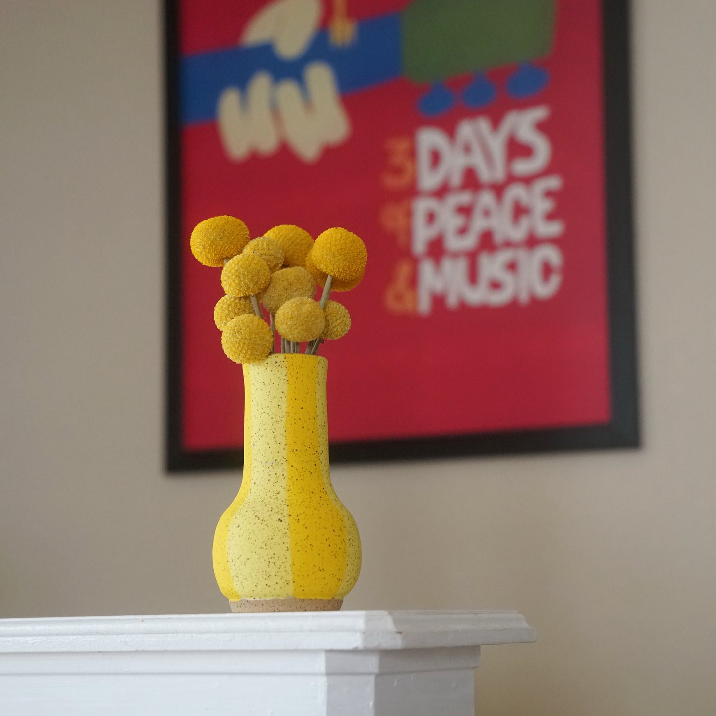 Yellow vase with decorative elements on a white mantel against a wall with a colorful poster.