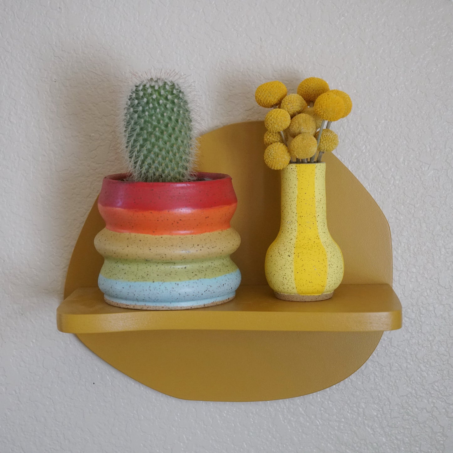 Round wooden shelf with colorful ceramic pots containing a cactus and yellow flowers on a plain wall.